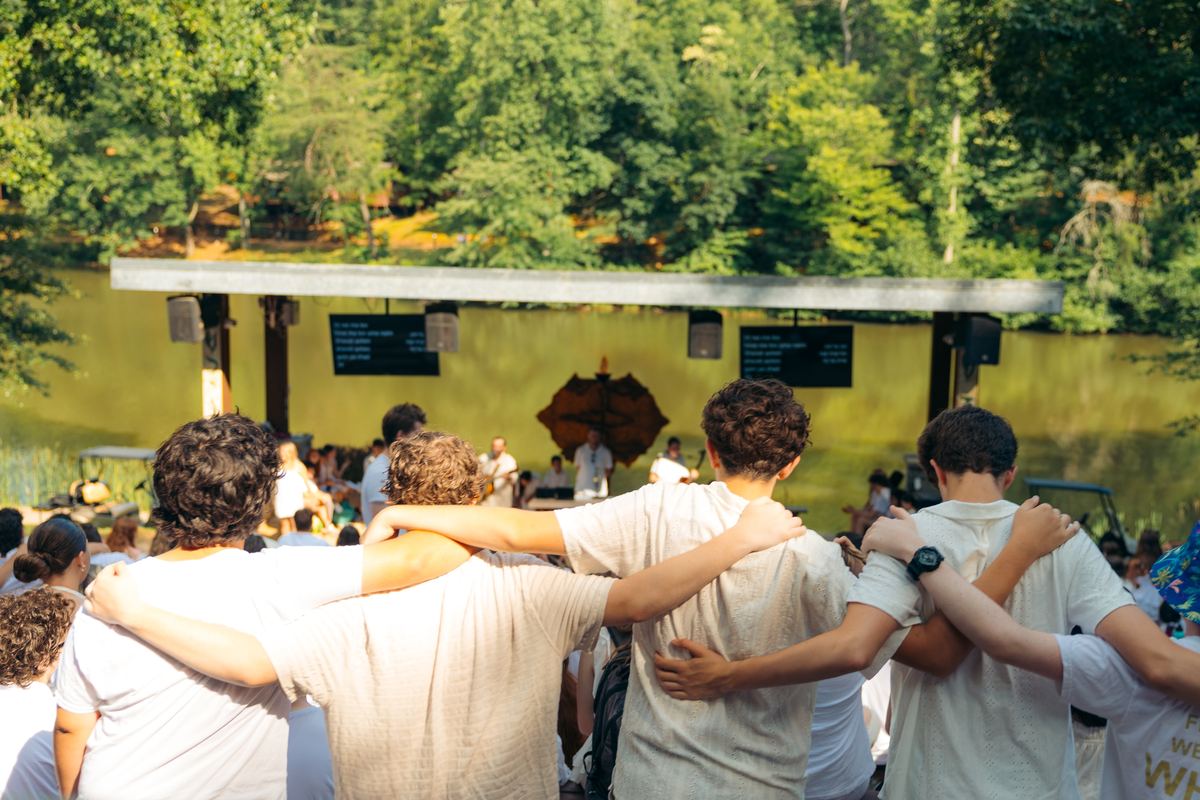 Campers in Shabbat white from behind, arms linked, facing a stage by the lake