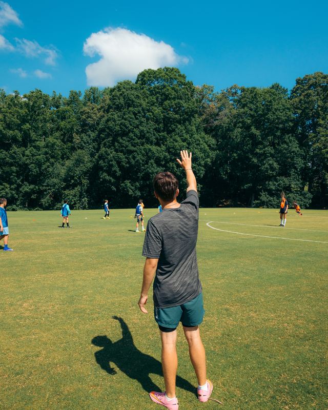 A counselor seen from behind, waving to campers across a sports field